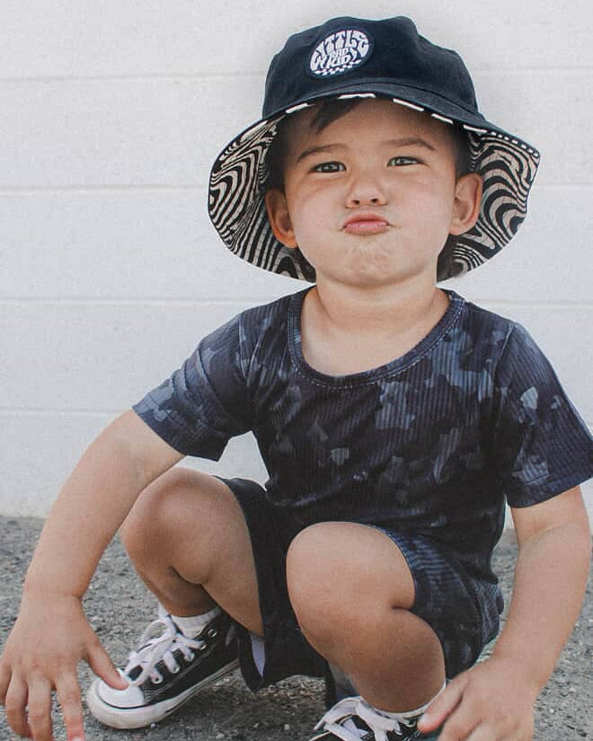 Child wearing a black bucket hat and black and white sneakers, squatting on a concrete surface.