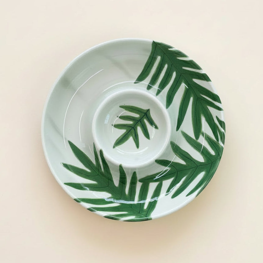 A white ceramic serving bowl with a matching saucer, decorated with a dark green hand-painted palm leaf pattern, set against a solid pale beige background.