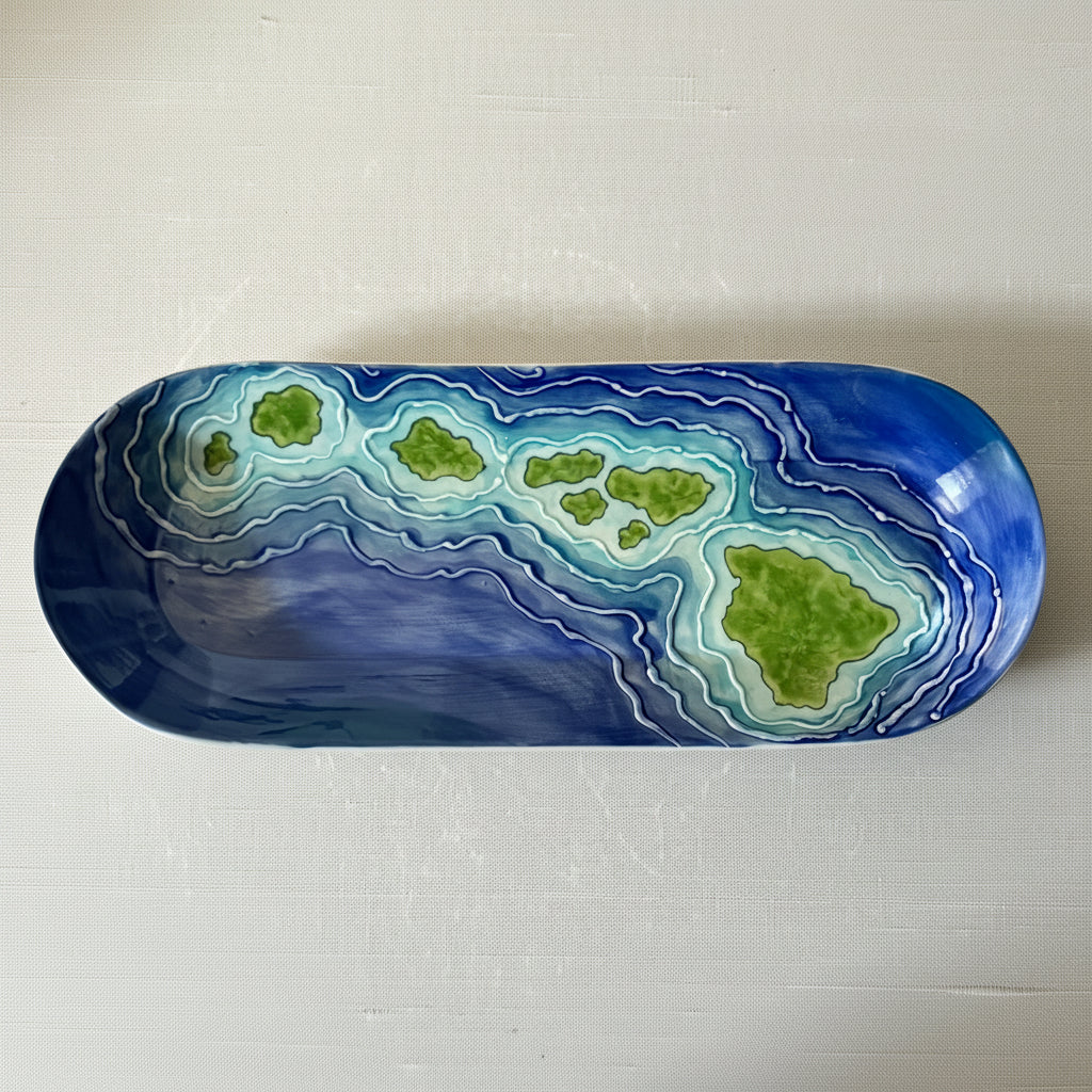 An oval ceramic plate with a hand-painted topographic design of green islands surrounded by concentric blue and white water-like patterns, set against a light textured background.