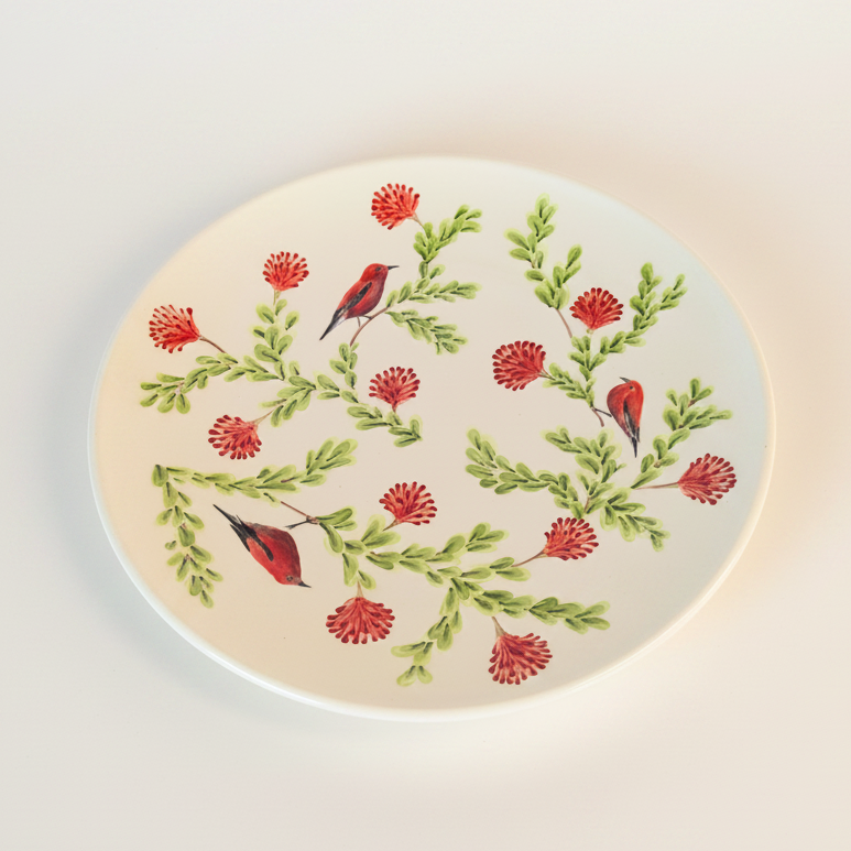 A hand-painted ceramic salad plate featuring a floral pattern with red and green colors and the image of a small, crimson Hawaiian honeycreeper bird.