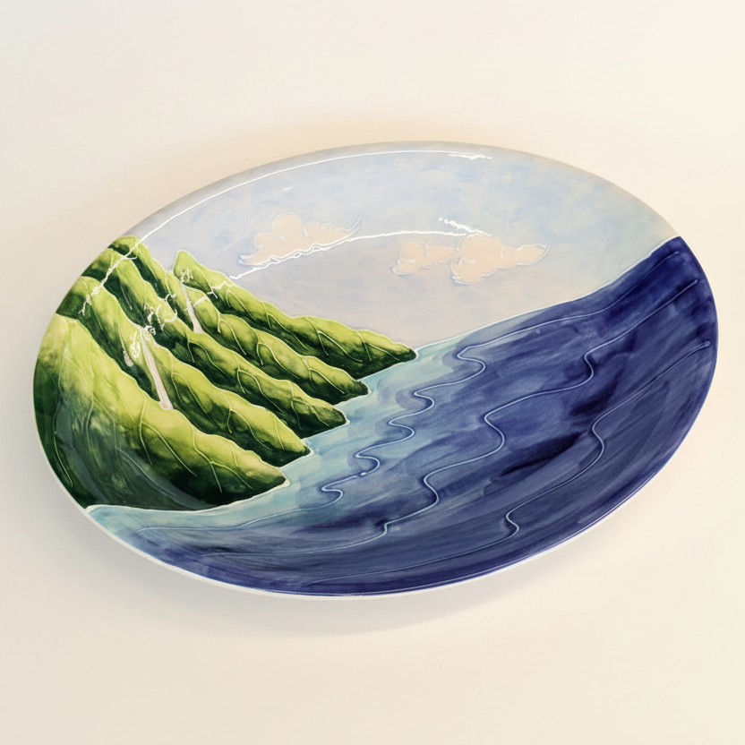 An oval-shaped decorative platter featuring a hand-painted design with a green and blue color scheme, portraying a stylized representation of a coastal landscape, specifically the Nā Pali Coast of Kauai.