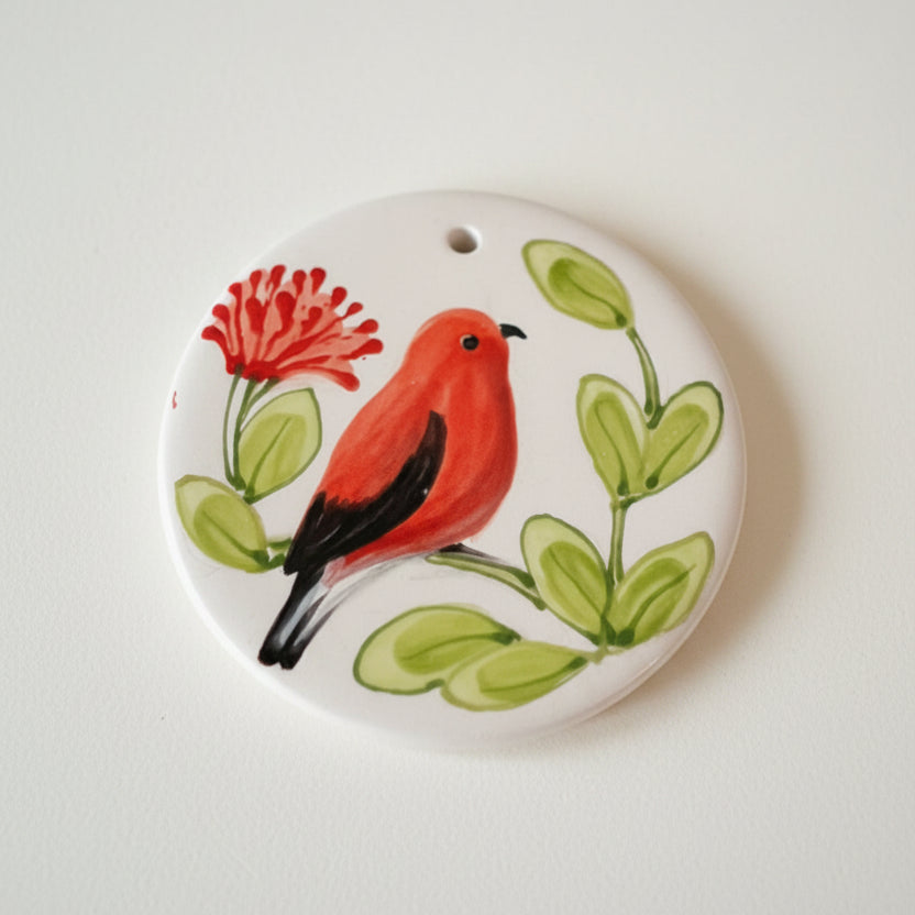 A round white ceramic ornament featuring a painted red bird with black wings, green leaves, and a red flower, shown against a plain light background.