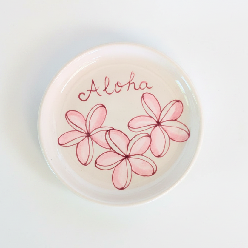 A round white ceramic dish with a hand-painted pink plumeria flower design and the word 'Aloha' in pink script, shown from a top-down view on a white background.