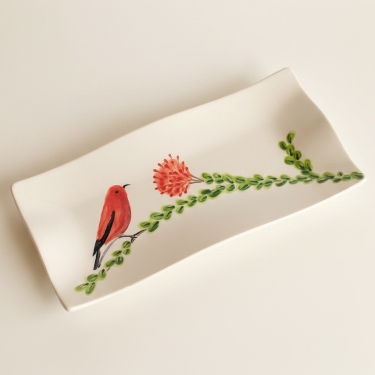 A white ceramic rectangular serving tray with a curved, wavy edge, decorated with a painted red bird perched on a green leafy branch next to a small pink flower, set against a plain light beige background.