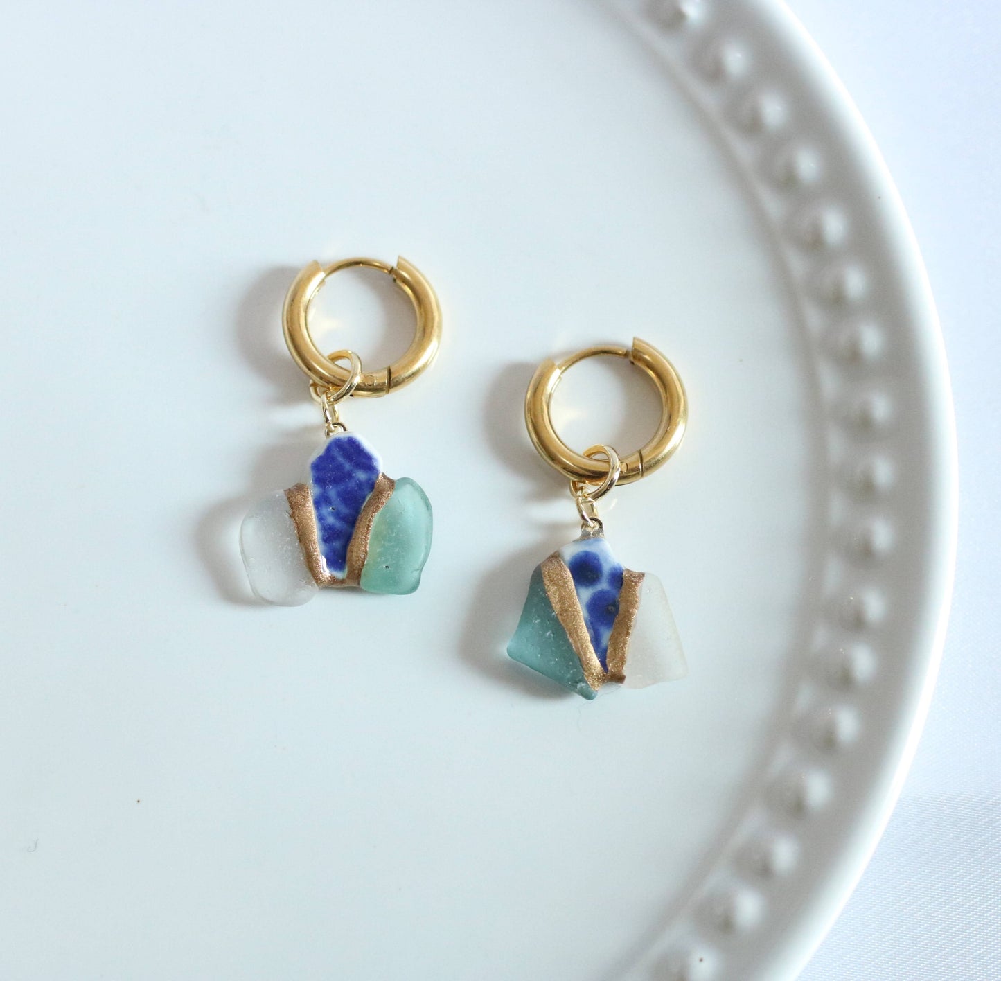 Gold hoop earrings with blue and green glass charms on a white plate.