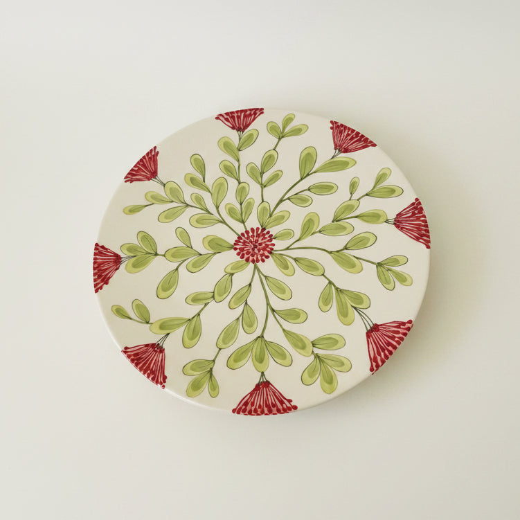 A round ceramic cake plate with a hand-painted botanical design featuring green leaves and red flower accents on a cream background, photographed from directly above on a light neutral surface.