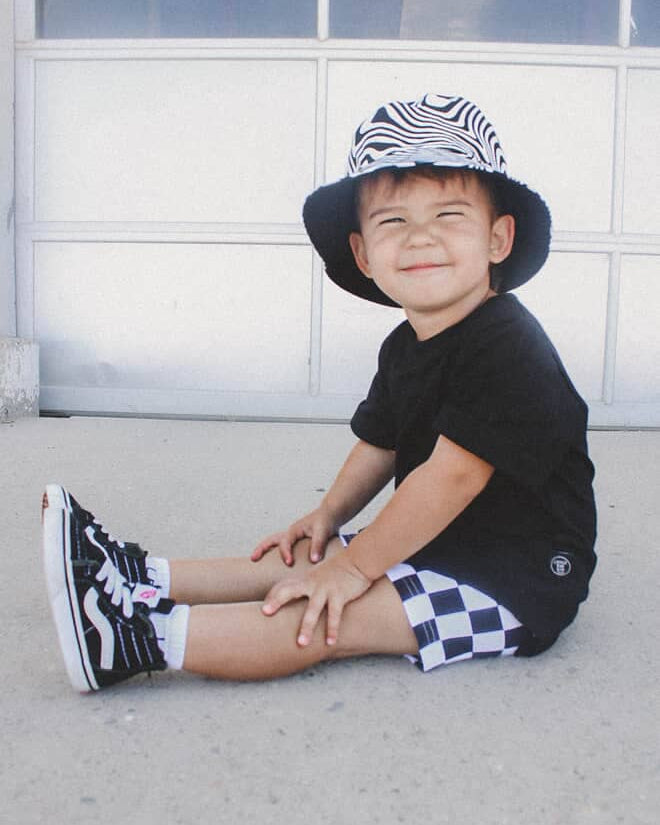 Child wearing a black and white checkered hat, black shirt, and checkered shorts sitting on the ground.