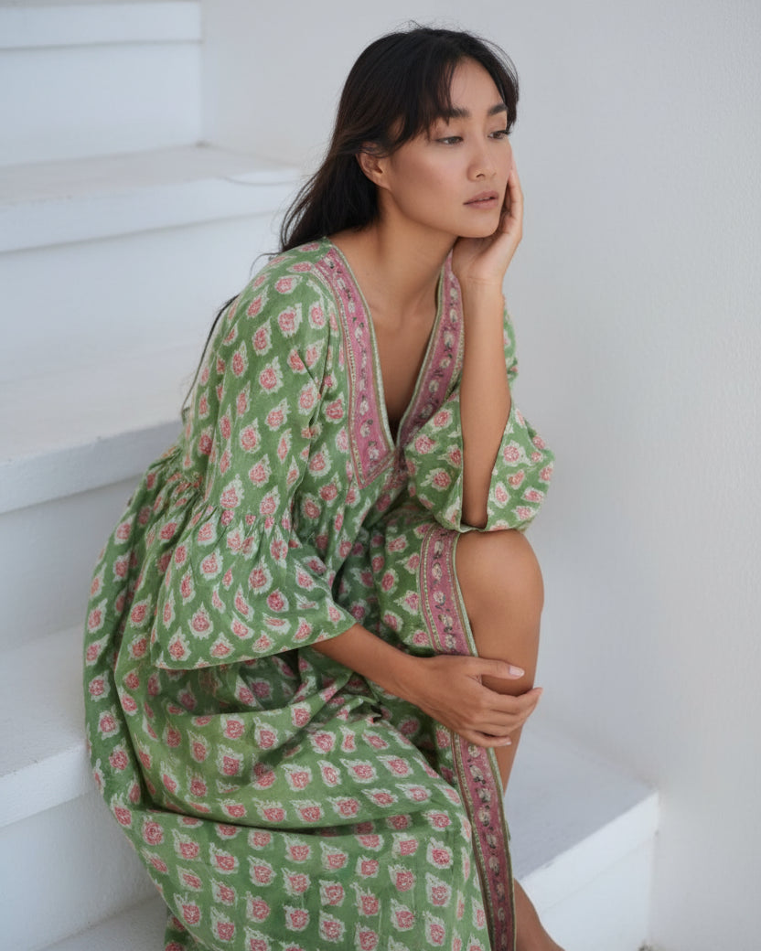 A woman wearing a green and beige floral print dress with three-quarter sleeves and a V-neckline, sitting in a relaxed pose with her hand near her face.