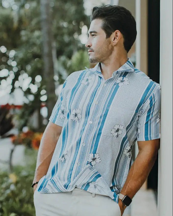 Man wearing a patterned aloha shirt standing outdoors with greenery in the background