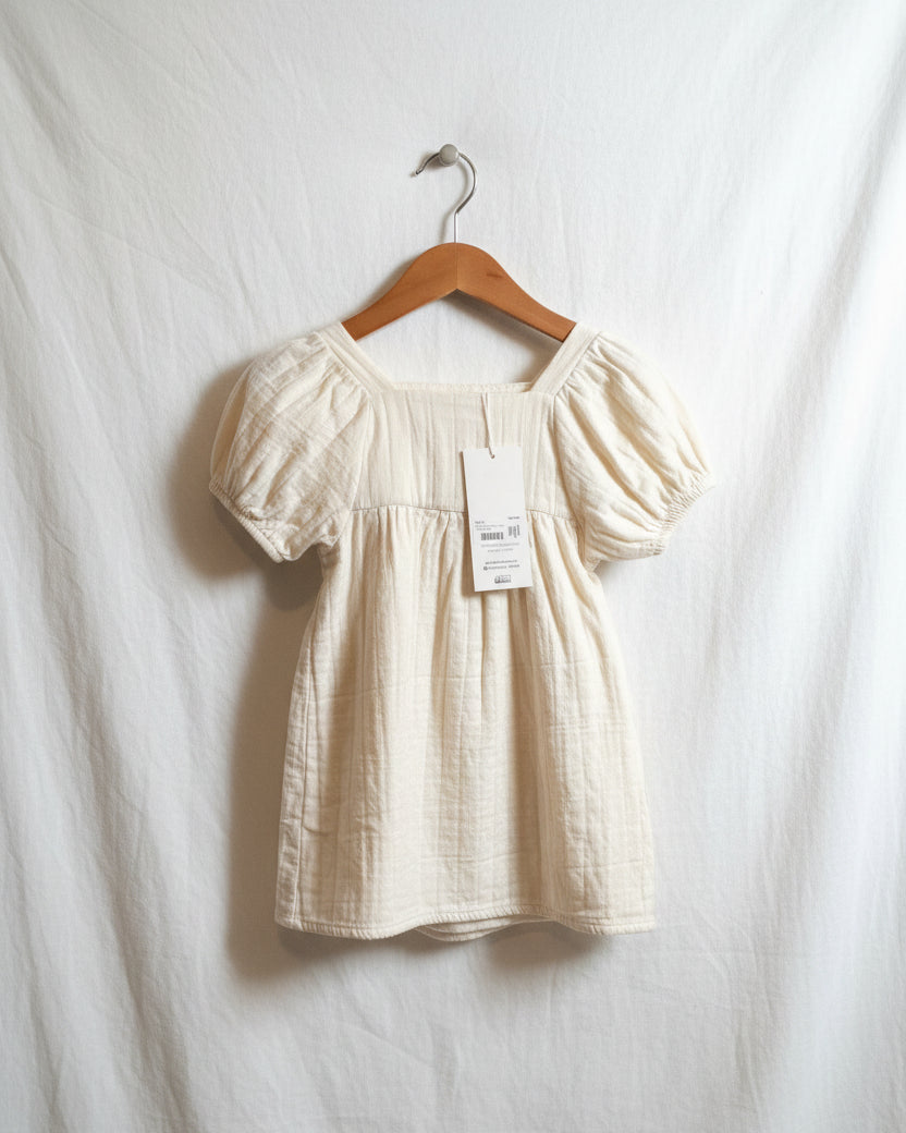 A white, short-sleeved baby girl's summer dress hanging on a wooden hanger in a wardrobe, with a tag attached to it.