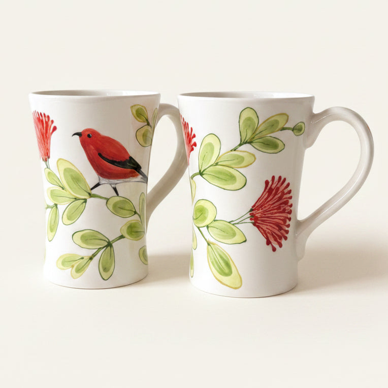 Two white ceramic mugs with a botanical and bird pattern, featuring red flowers and green leaves with a red bird, set against a plain light beige background.