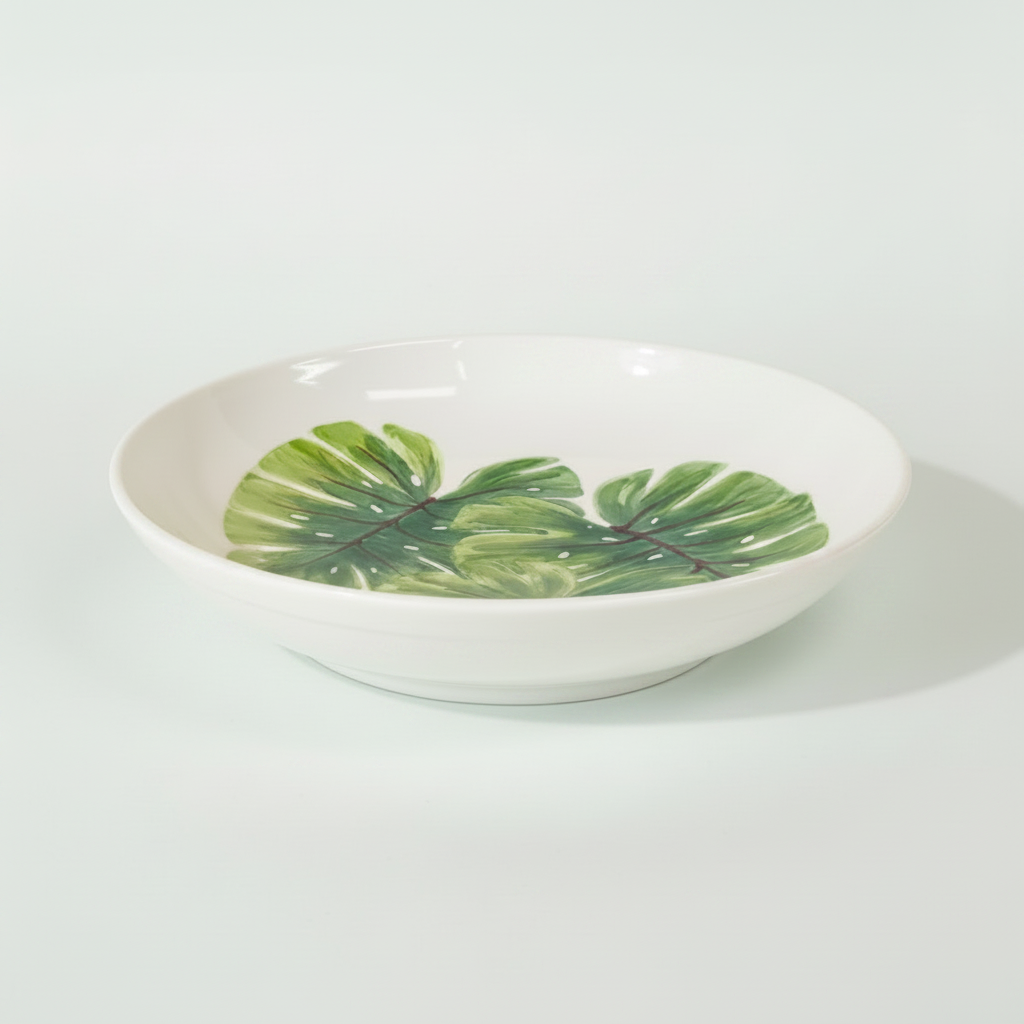 A white ceramic shallow bowl decorated with a painted green monstera leaf pattern, set against a solid light beige background.