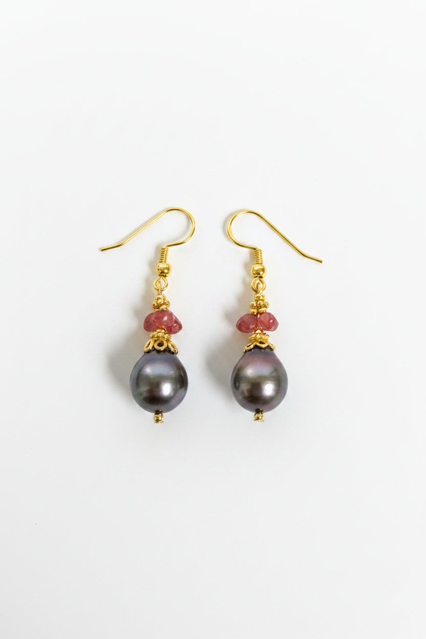 A pair of gold dangle earrings featuring large, dark gray-black freshwater pearl drops and small red faceted beads, shown on a plain off-white background.