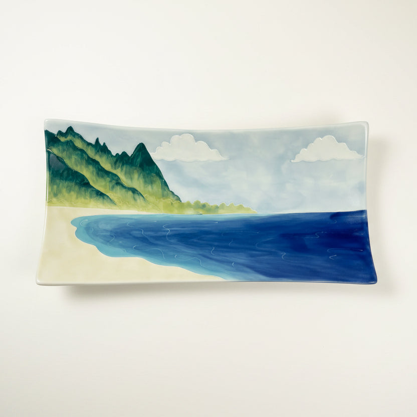A rectangular ceramic serving platter featuring a hand-painted coastal landscape with green mountains, a sandy beach, blue water, and white clouds, shown against a plain white background.