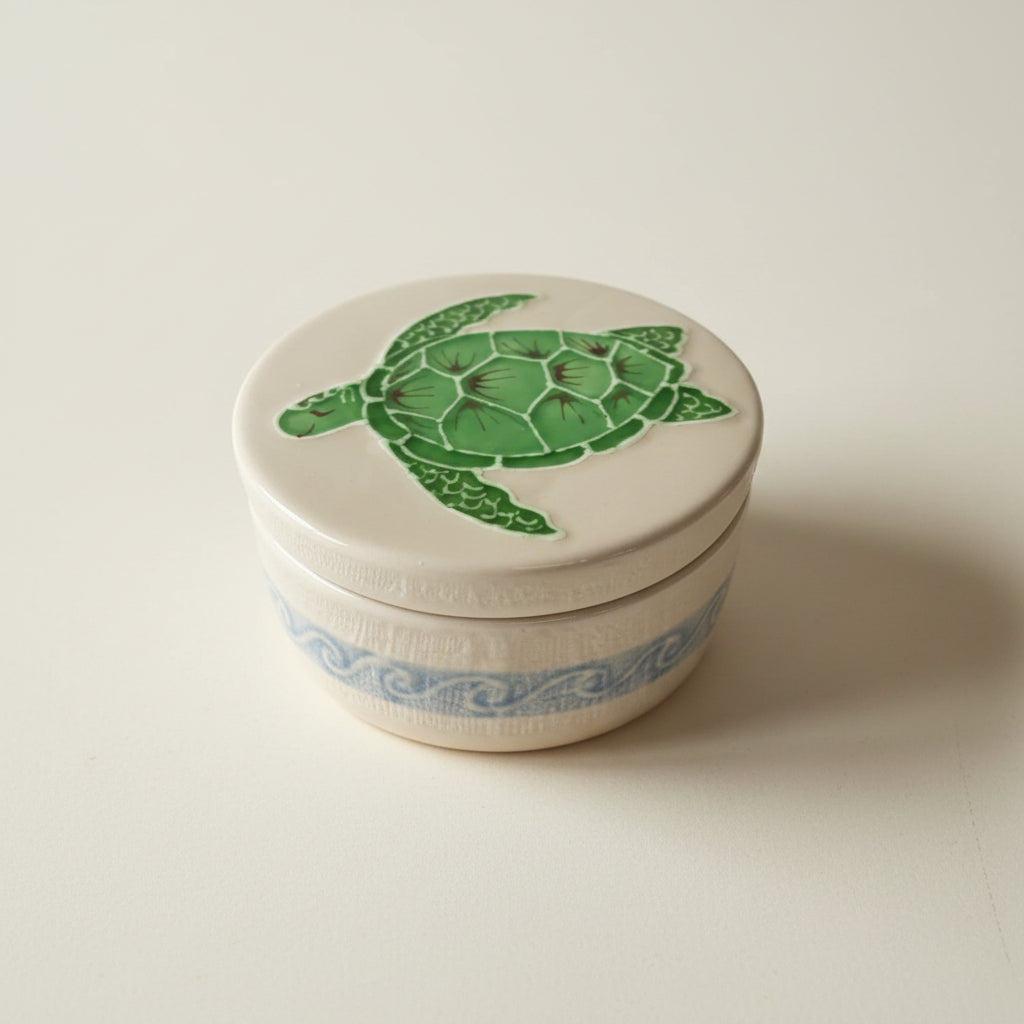 A beige ceramic treasure box with a green turtle design on the lid, placed on a white textured surface.
