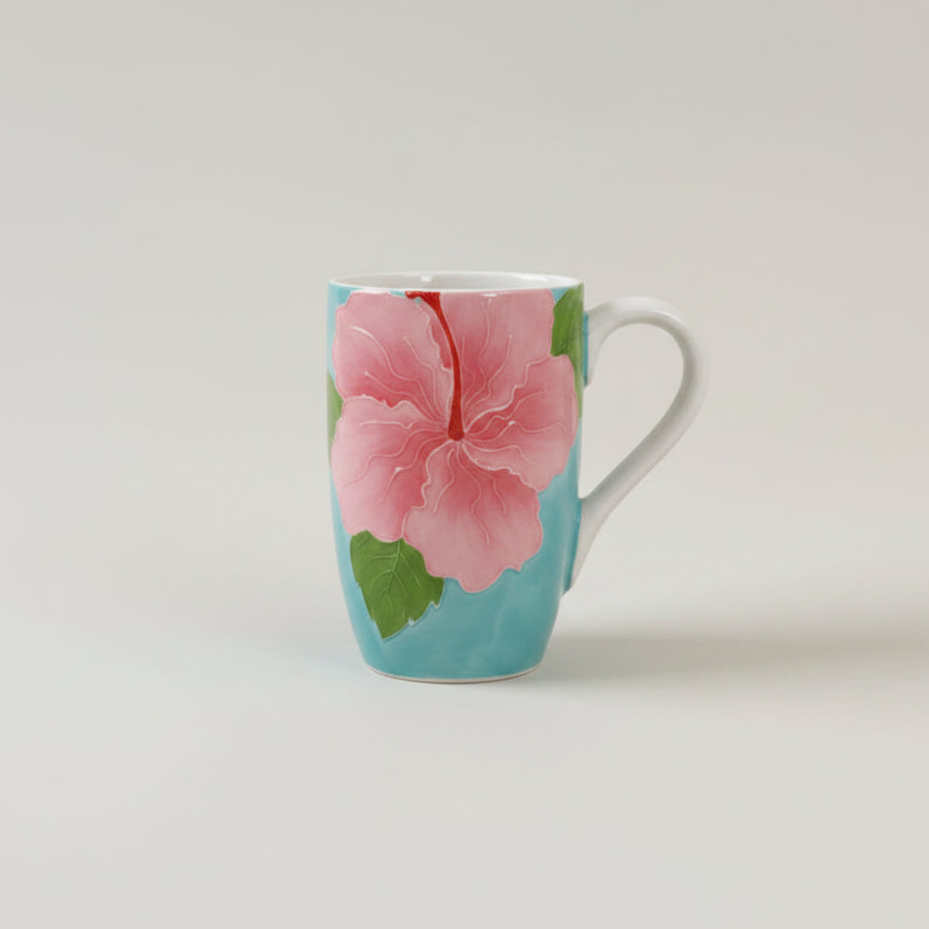 ceramic mug with white handle and interiors, decorated with large pink hibiscus flower and green leaves on a turquoise background, shown on a light gray studio background.