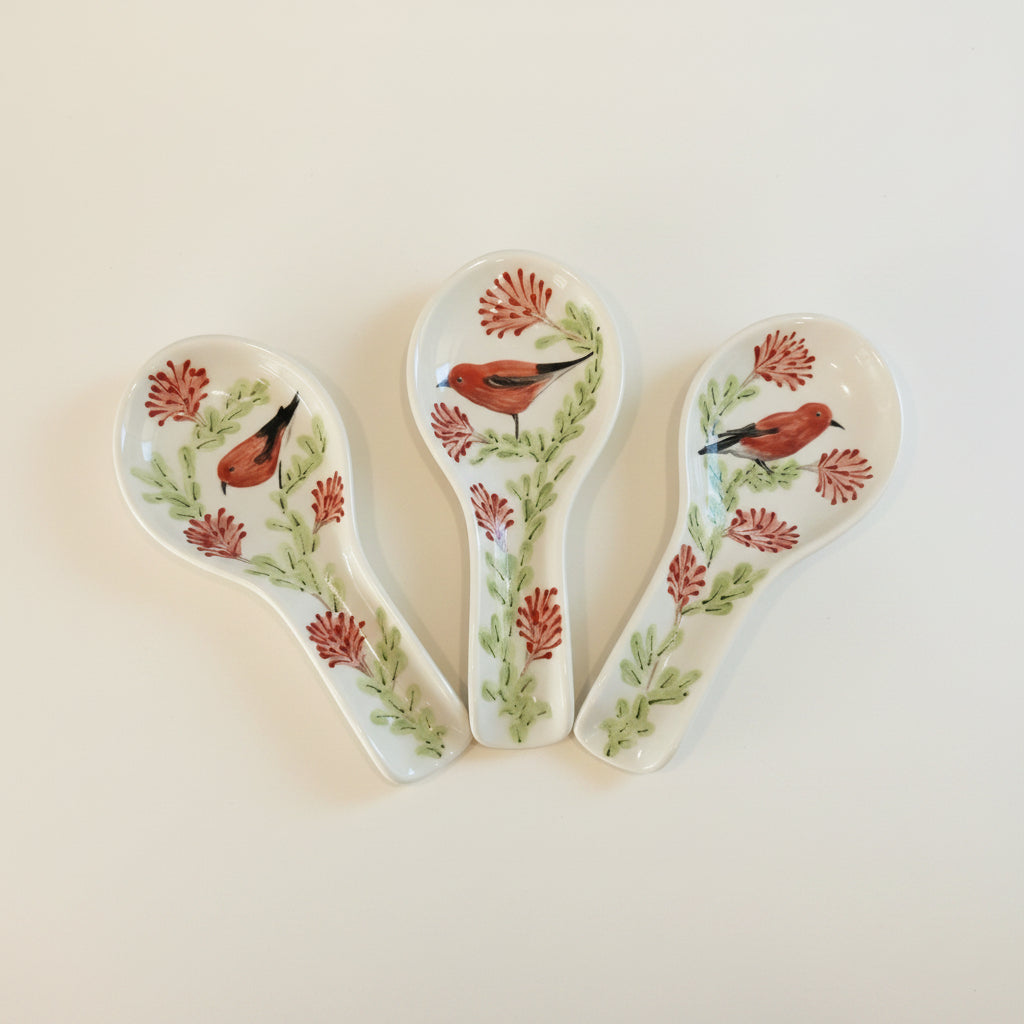 Three white ceramic soup spoons decorated with painted red birds, green leaves, and red flowers, arranged side by side on a light background.