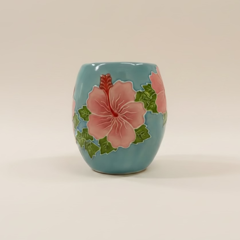A small ceramic cup with a rounded shape, decorated with a pink hibiscus flower and green leaves on a light blue-green glazed background, set against a plain light beige studio backdrop.