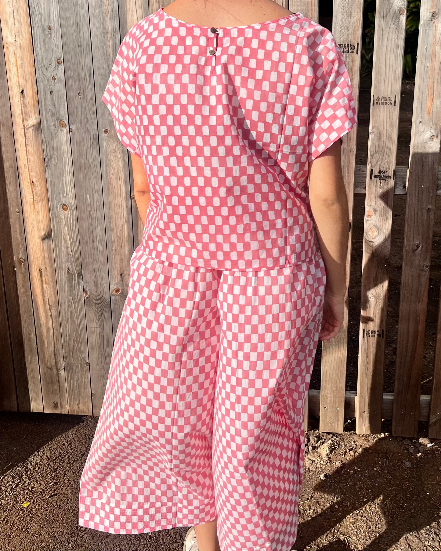 Woman wearing a pink checkered top and pants standing in front of a wooden fence.