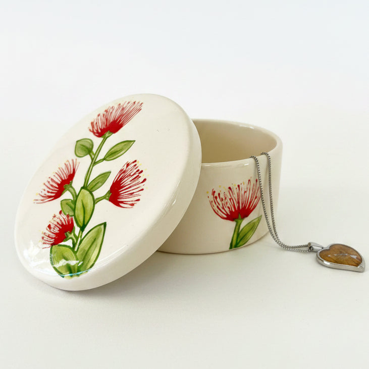 An off-white ceramic keepsake box with a matching lid, decorated with red flowers and green leaves, shown with a silver heart-shaped pendant necklace on a white background.
