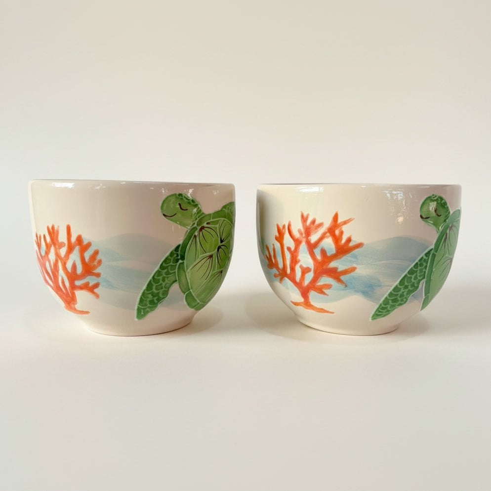 Two small white ceramic cups with painted sea turtles and orange coral, shown side by side on a plain light background.