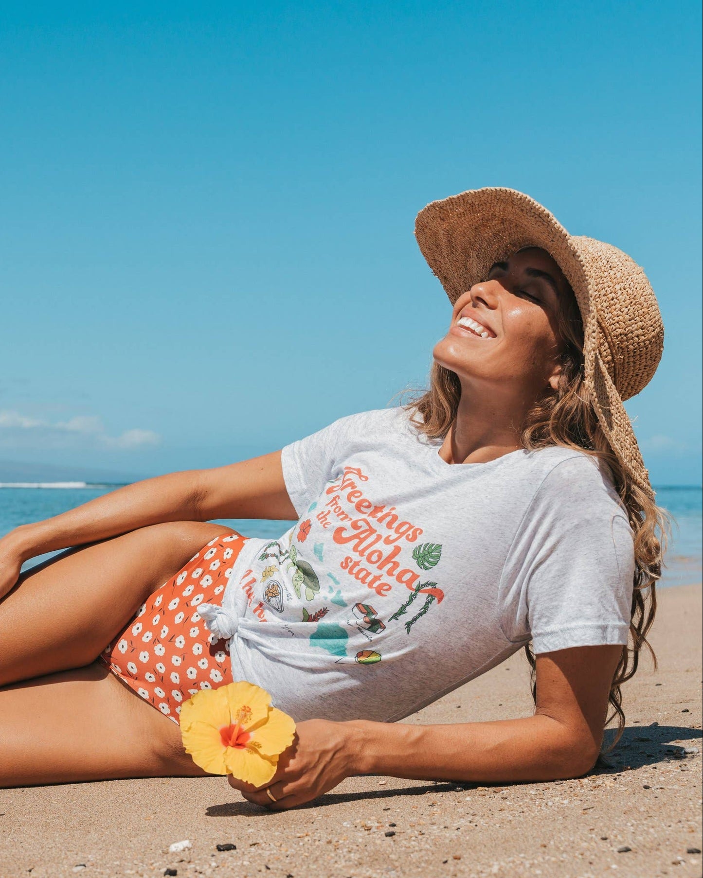 Woman sitting on a beach wearing a straw hat and colorful outfit, holding a flower.