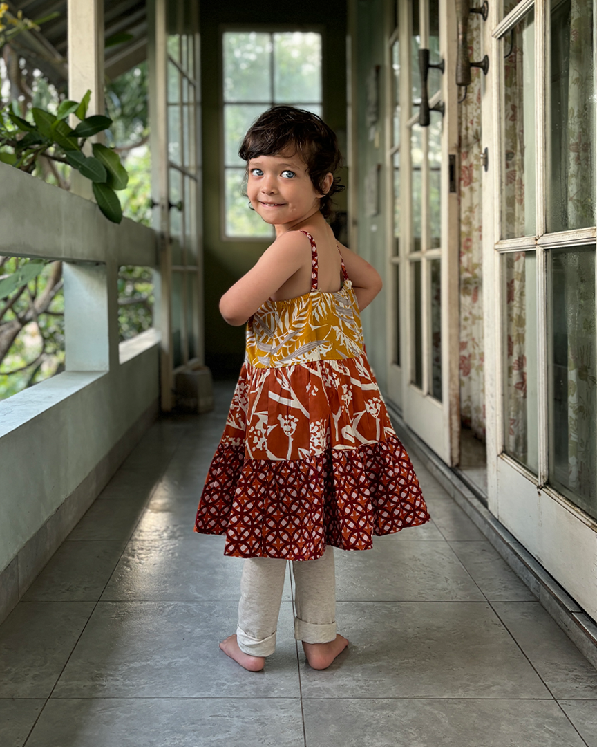 Child wearing a patterned dress standing on a tiled floor with greenery outside.