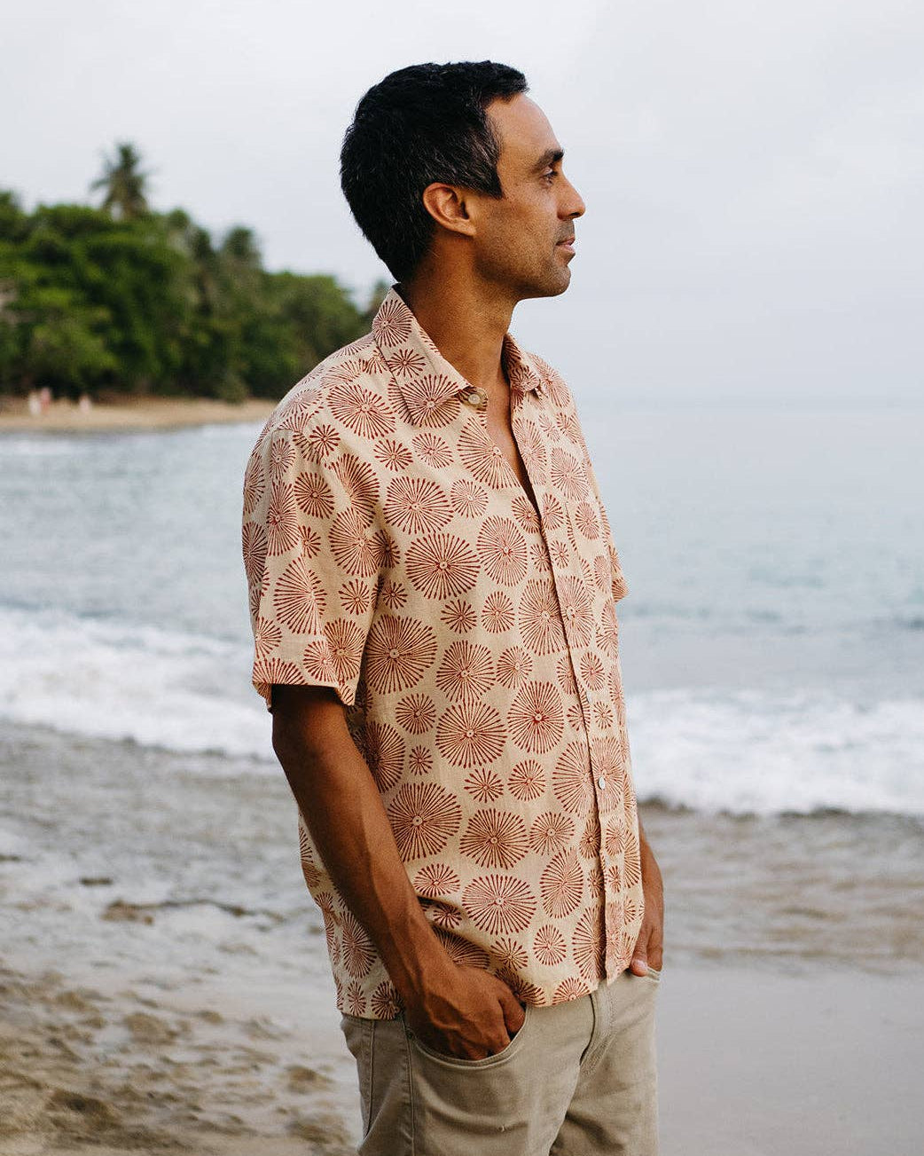 Man wearing a patterned aloha shirt standing on a beach with ocean and trees in the background