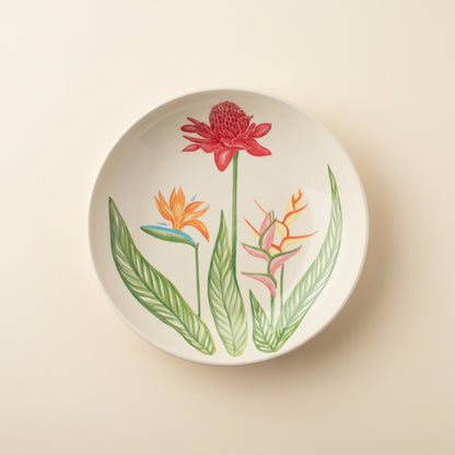 A round white ceramic plate featuring a botanical illustration of tropical flowers, including a red ginger flower, an orange bird of paradise, and pink heliamphora flowers with green leaves, shown on a clean white background.