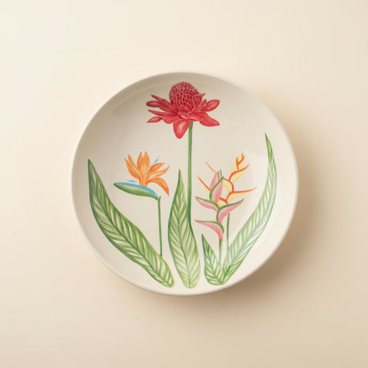 A round white ceramic plate featuring a botanical illustration of tropical flowers, including a red ginger flower, an orange bird of paradise, and pink heliamphora flowers with green leaves, shown on a clean white background.