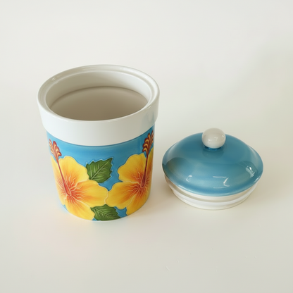 A ceramic canister with a matching lid, decorated with yellow hibiscus flowers and green leaves on a blue background, set against a plain off-white studio backdrop.