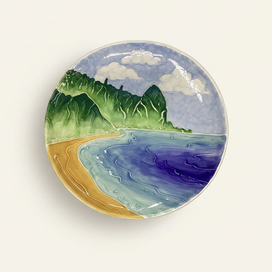 A round ceramic dinner plate with a hand-painted design featuring a coastal landscape with a beach, ocean, and hills.