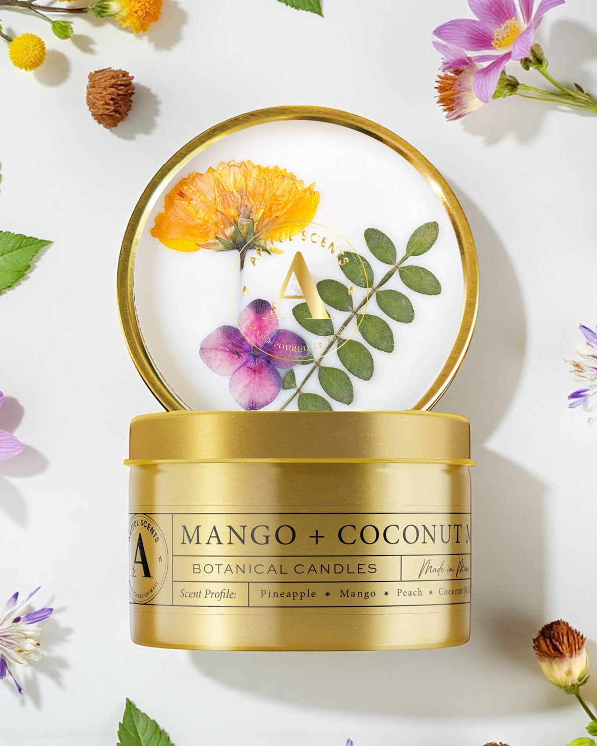 Gold candle container labeled 'Mango + Coconut' surrounded by flowers and leaves on a white background