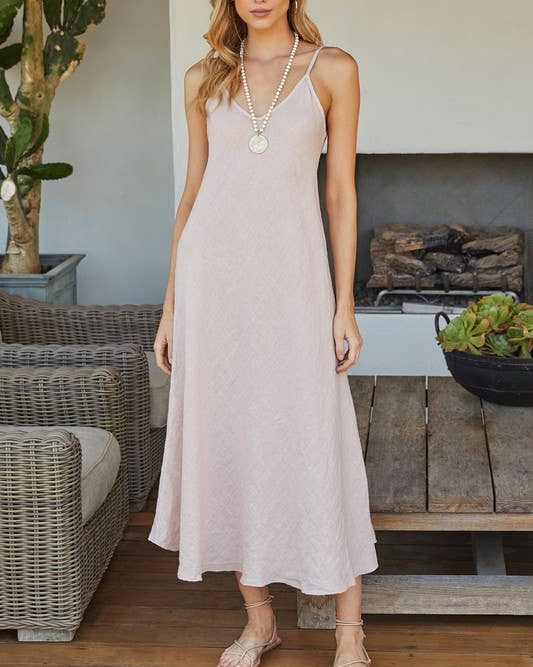 Woman in a light pink dress standing in an outdoor setting with plants and a fireplace.