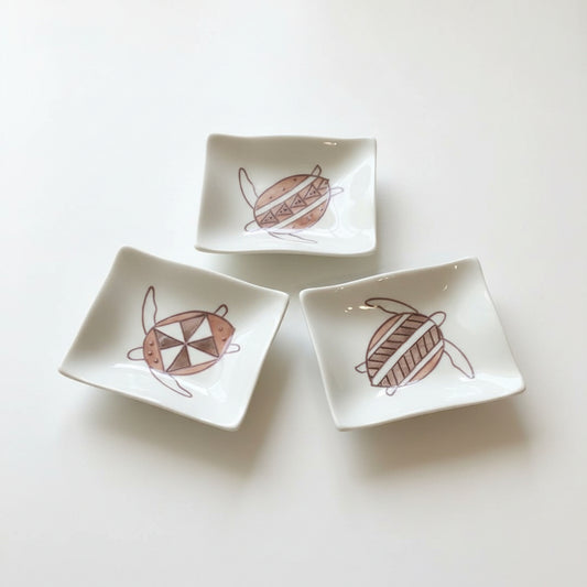 Three small white square ceramic dishes with rounded corners, each decorated with a brown turtle illustration, arranged on a wooden surface with a woven mat and a black plate with a white mug partially visible.