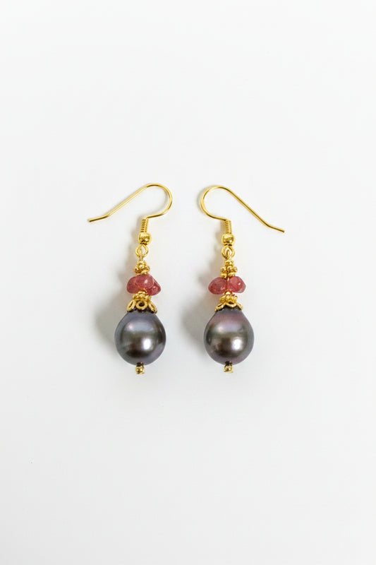 A pair of gold dangle earrings featuring large, dark gray-black freshwater pearl drops and small red faceted beads, shown on a plain off-white background.