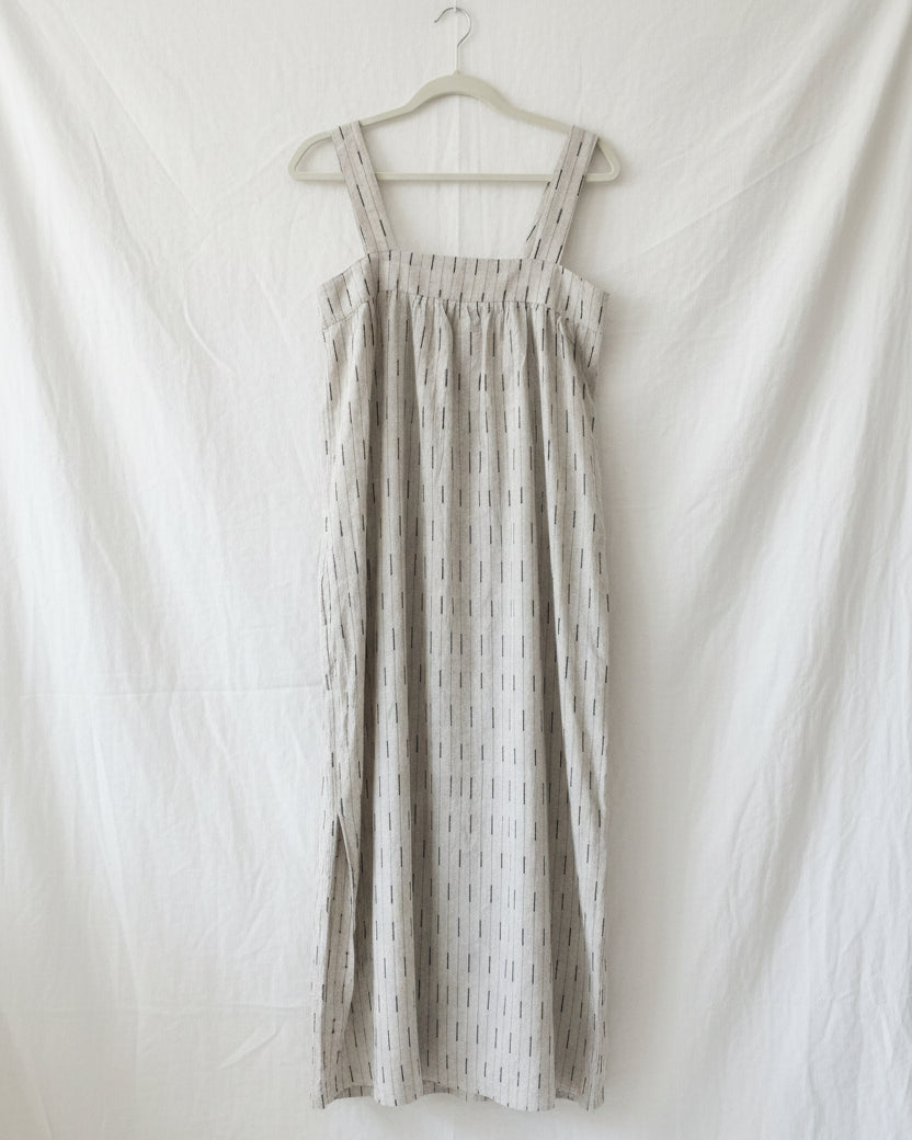 A white embroidered linen dress displayed on a hanger, featuring a gathered waist and sleeveless design.