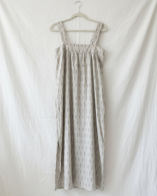 A white embroidered linen dress displayed on a hanger, featuring a gathered waist and sleeveless design.