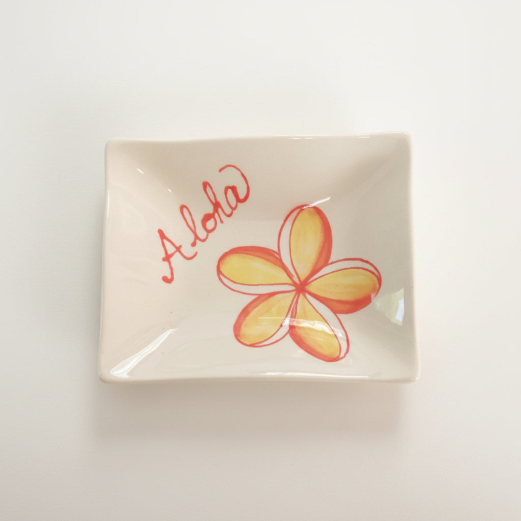 A white ceramic sauce dish with a red 'Aloha' greeting and a plumeria flower design in orange and yellow.