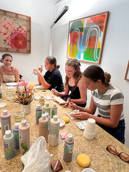 Three women standing around a table with various bottles and items, with colorful abstract art on the wall.