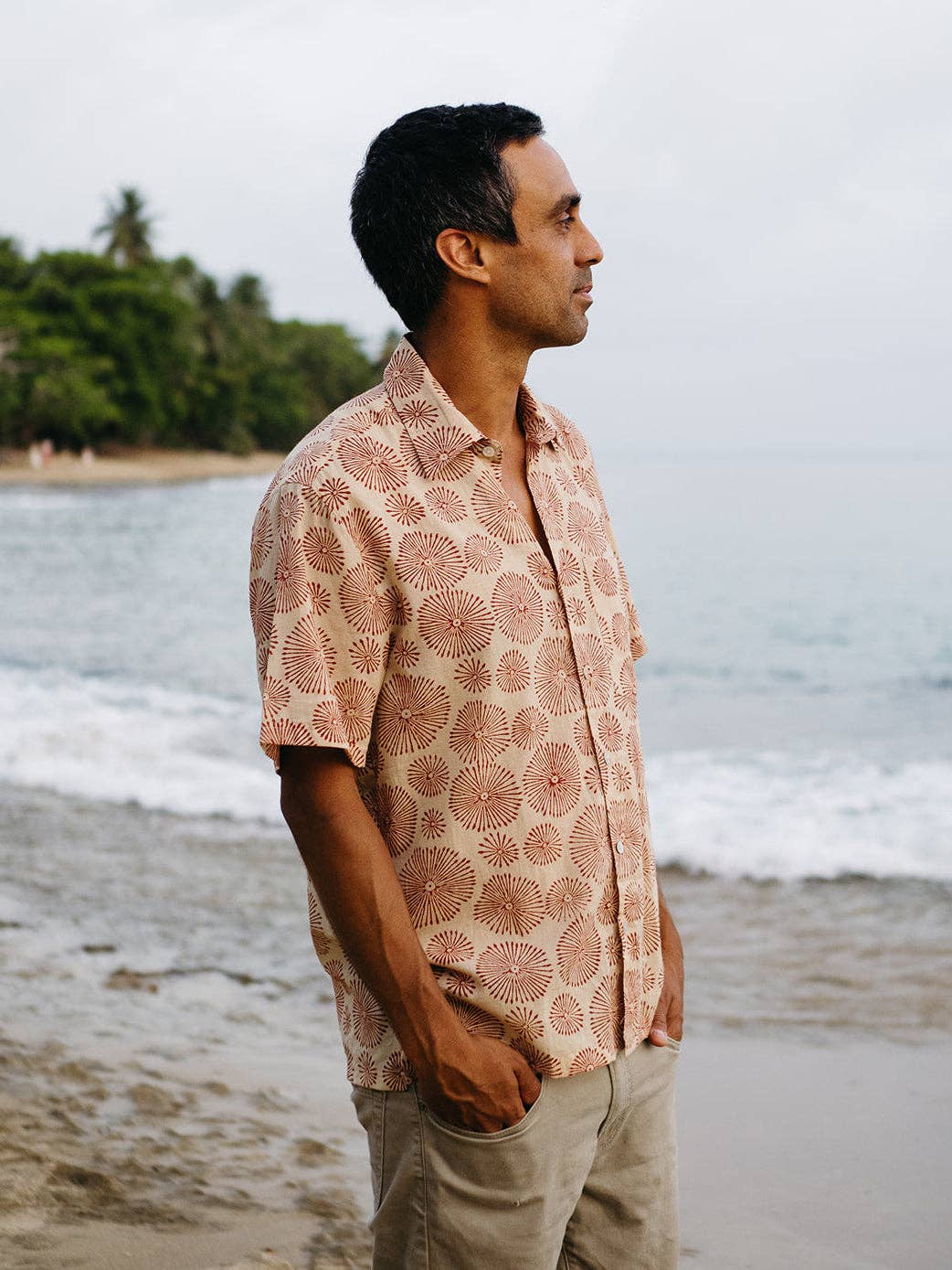 Man wearing a patterned shirt standing on a beach with ocean and trees in the background