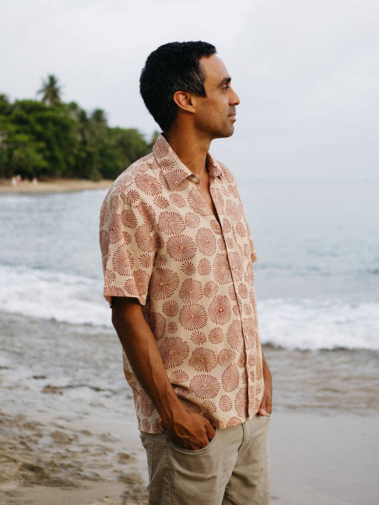 Man wearing a patterned shirt standing on a beach with ocean and trees in the background